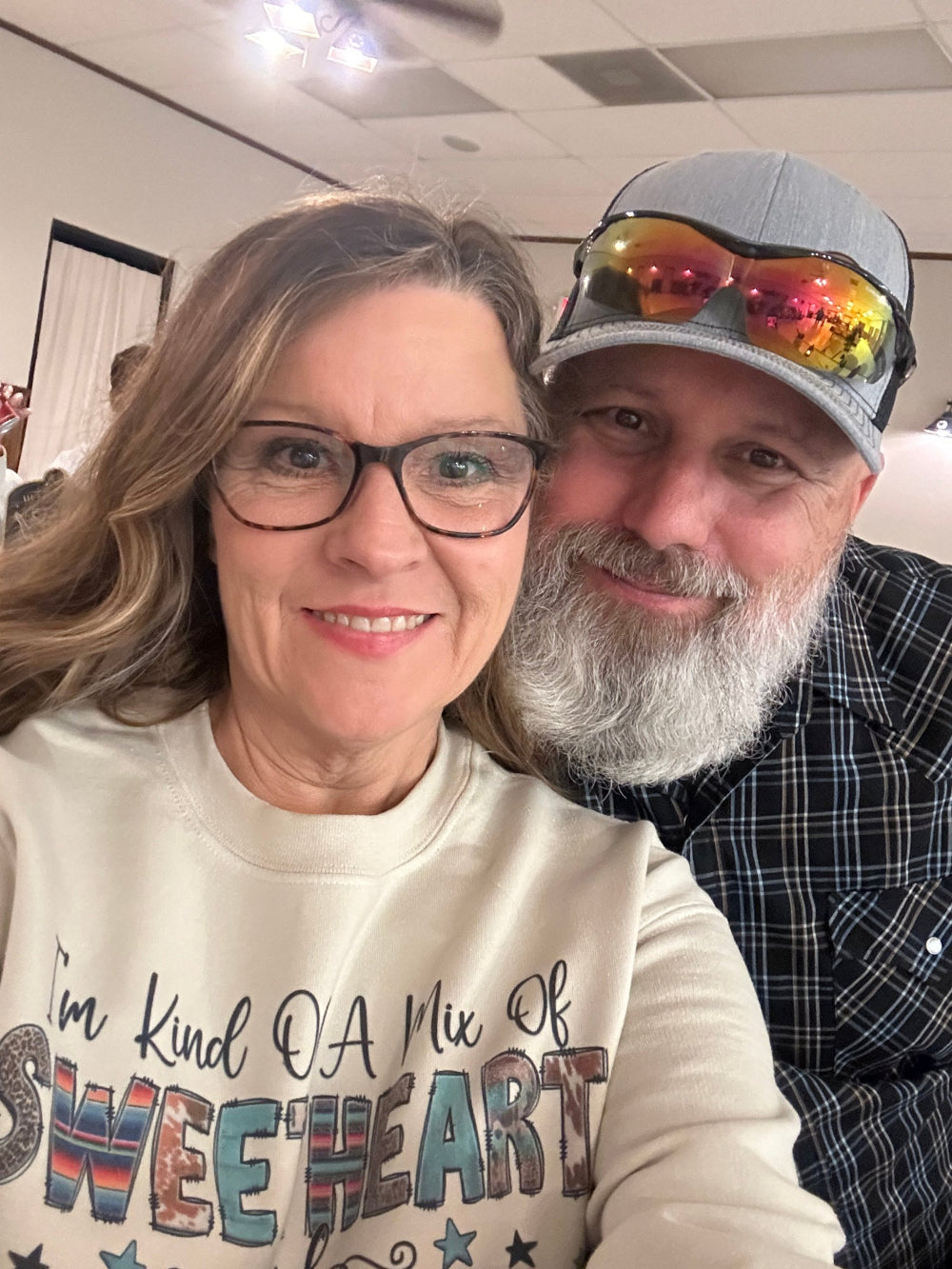 Two people taking a selfie indoors, one wearing a colorful cap and plaid shirt, the other in a light-colored sweatshirt with text. Roger and Brenda Nicholson, owners of Lula Belle Creations.